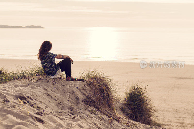 一名女子坐在沙丘上，望着日落时分康沃尔郡的Gwithian Towans海滩。